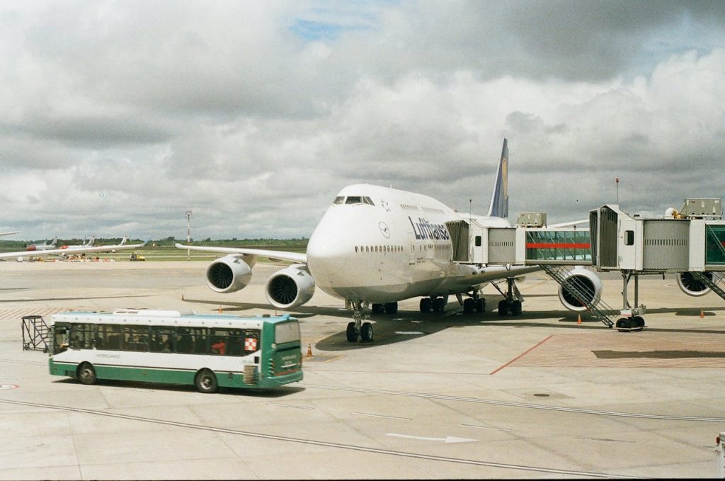 A large airplane docked at an airport gate with a shuttle bus nearby under cloudy skies.
